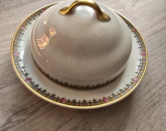 May include: A white porcelain butter dish with a gold handle and a matching plate. The plate has a gold rim and a decorative floral pattern with pink and green accents. The dish is designed to hold butter.