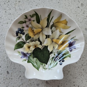 May include: A white ceramic dish shaped like a seashell with a floral design. The design features yellow daffodils, purple and white flowers, green leaves, and blue berries.