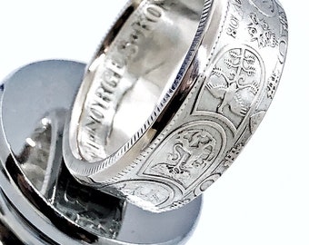 May include: A silver-toned coin ring featuring intricate embossed details on the exterior band, including a bird motif and the text 1918. The interior of the band displays the engraved text NORGES KONGE, showcasing a polished metallic finish.