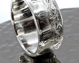 May include: A silver-toned ring with a wide band featuring intricate embossed scrollwork patterns and the letters M, O, R, and I clearly visible along the outer surface. The metal has a polished, reflective finish with a smooth interior.
