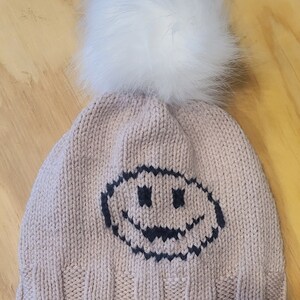 May include: A knitted beanie hat in a light pink color with a white faux fur pom pom on top. The hat has a black embroidered smiley face on the front.