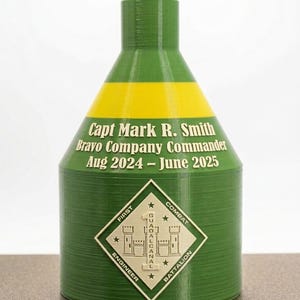 May include: A green, cone-shaped object with a yellow horizontal stripe. It features the text: Capt Mark R. Smith, Bravo Company Commander, Aug 2024 – June 2025. Below is a diamond-shaped emblem with the text First Combat Engineer Battalion.