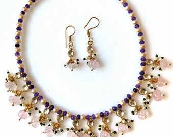 Puede incluir: Un collar de oro con cuentas moradas y crema y dijes en forma de flor rosa. Aretes a juego con dijes en forma de flor rosa.