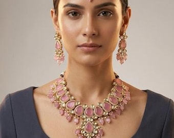 Puede incluir: Un conjunto de joyería que incluye un collar dorado, pendientes colgantes a juego y un maang tikka. Cada pieza está adornada con piedras rosas en forma de lágrima y pequeños cristales transparentes, dispuestos en un patrón floral con cuentas rosas colgantes.
