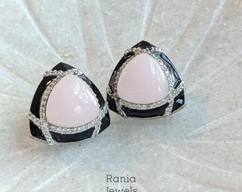 Puede incluir: Un par de pendientes de botón triangulares. Cada pendiente presenta una piedra central de color rosa claro, enmarcada por un borde de esmalte negro e incrustada con pequeños cristales brillantes. Los pendientes están sobre un fondo neutro.