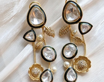 Puede incluir: Un par de pendientes dorados con piedras claras y facetadas y ribete de esmalte negro. Los pendientes presentan detalles florales y de hojas, con piedras colgantes en forma de lágrima y ovaladas, y pequeñas perlas de imitación.