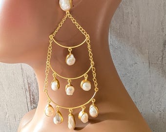 Puede incluir: Pendientes colgantes dorados con múltiples niveles. Cada nivel presenta una serie de perlas blancas de forma irregular. Los pendientes están diseñados con un efecto en cascada, creando un aspecto dramático y elegante. Los pendientes están sujetos a un maniquí.