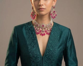 Puede incluir: Una chaqueta de color verde azulado profundo con bordados tonales de pavos reales a lo largo de las solapas. El conjunto se complementa con un collar de perlas de varias hileras, pendientes a juego y un adorno frontal con piedras rosas.