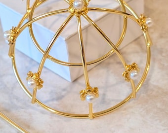 Puede incluir: Un accesorio para el cabello de metal dorado con un diseño de jaula esférica adornado con pequeñas perlas blancas en cada intersección. La estructura consta de varillas metálicas curvas y un palillo para el cabello a juego con punta de perla.