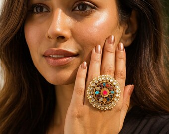 Puede incluir: Un anillo grande y circular de tono dorado con una piedra central rosa rodeada por un patrón floral de piedras más pequeñas en azul, rojo y transparente. El borde exterior está adornado con una hilera de pequeñas cuentas blancas redondas.
