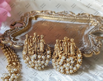 Puede incluir: Un par de pendientes dorados con un relieve detallado de tres figuras sobre un grupo de pequeñas perlas blancas. Los pendientes tienen una base circular y un acabado texturizado, mostrando una artesanía y un diseño intrincados.