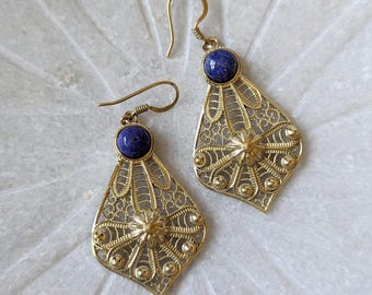 Puede incluir: Un par de pendientes de filigrana dorada con detalles de piedras preciosas azules. Los pendientes tienen forma de lágrima y un diseño delicado e intrincado.