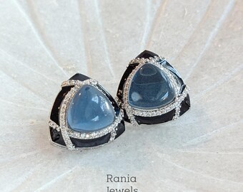 Puede incluir: Un par de pendientes de botón triangulares. Cada pendiente presenta una piedra preciosa azul claro engastada en un marco negro y plateado, acentuada con pequeños cristales transparentes. Los pendientes son de Rania Jewels.