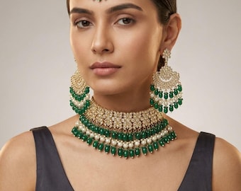 Puede incluir: Un conjunto de joyería en tono dorado que incluye un collar tipo gargantilla, pendientes largos y un maang tikka. Cada pieza está adornada con piedras transparentes, perlas blancas y cuentas verdes en forma de lágrima que cuelgan.