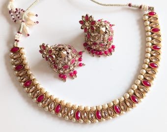 Puede incluir: Un conjunto de joyería que consta de un collar y dos pendientes jhumka. El collar presenta una alternancia de piedras fucsia de talla marquesa y cristales transparentes, bordeados por pequeñas perlas blancas. Los pendientes a juego tienen.