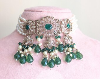 Puede incluir: Un collar gargantilla plateado con un diseño floral, con detalles en verde esmeralda y blanco. El collar tiene múltiples hilos de cuentas blancas y gemas verdes colgantes en forma de lágrima. El texto "Rania Jewels" es visible.