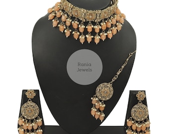 Puede incluir: Un conjunto de joyas de novia indias de color melocotón con un collar choker, pendientes y un maang tikka. El conjunto presenta metal dorado con intrincados diseños florales y cuentas de color melocotón.