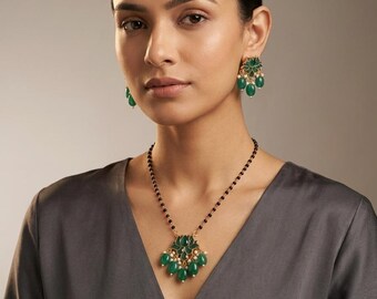 Puede incluir: Un collar de tono dorado con un colgante central de piedras verdes en forma de lágrima y pequeñas cuentas blancas, suspendido de una cadena de cuentas negras. Los pendientes a juego presentan un diseño floral similar en verde y blanco.