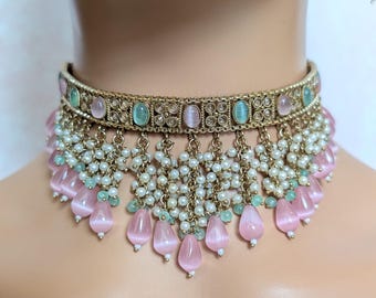 Puede incluir: Un collar gargantilla dorado con detalles en rosa y verde. El collar presenta piedras preciosas rosas y verdes de forma ovalada, pequeñas perlas blancas y cuentas rosas colgantes en forma de lágrima. El collar está diseñado para colocarse alrededor del cuello.