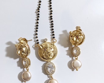 Puede incluir: Un collar de oro con un colgante de tigre y pendientes a juego. El colgante y los pendientes presentan un diseño de cabeza de tigre con una piedra roja para el ojo y piedras transparentes alrededor del borde. El collar está hecho de cuentas negras y los pendientes tienen una gota de perla.