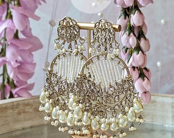Puede incluir: Un par de pendientes tipo candelabro en tono dorado con filigrana metálica, piedras transparentes y filas de cuentas blancas. Los pendientes están adornados con perlas colgantes en forma de lágrima color crema y pequeños detalles de cuentas, Rania Jewels.