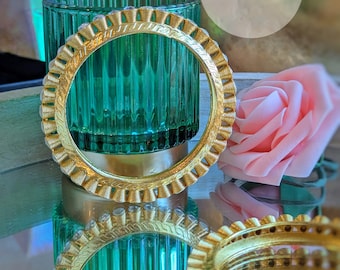 Puede incluir: Un brazalete dorado con un diseño decorativo y brillantes transparentes. El brazalete está apoyado en un portavelas de vidrio verde. La imagen también incluye una rosa rosa y el texto "Rania Jewels".