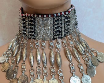Puede incluir: Collar gargantilla plateado con base de tela negra. El collar presenta intrincados trabajos en metal, colgantes con forma de moneda y colgantes alargados y decorativos. La banda superior de la gargantilla está adornada con cuentas rojas, blancas y negras.