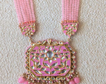 Puede incluir: Un collar de varias hebras con cuentas de color rosa claro y un gran colgante rectangular. El colgante tiene un acabado de esmalte rosa brillante con detalles en metal dorado y piedras transparentes. Una fila de cuentas en forma de gota cuelga.