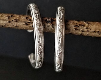 May include: Two silver bracelets with a floral design. The bracelets are on a wooden branch.