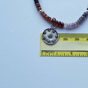 K&ouml;nnte beinhalten: Eine silberne Anh&auml;nger-Kette mit einem runden, silbernen Anh&auml;nger mit einem floralen Design. Die Kette besteht aus kleinen, mehrfarbigen Perlen.