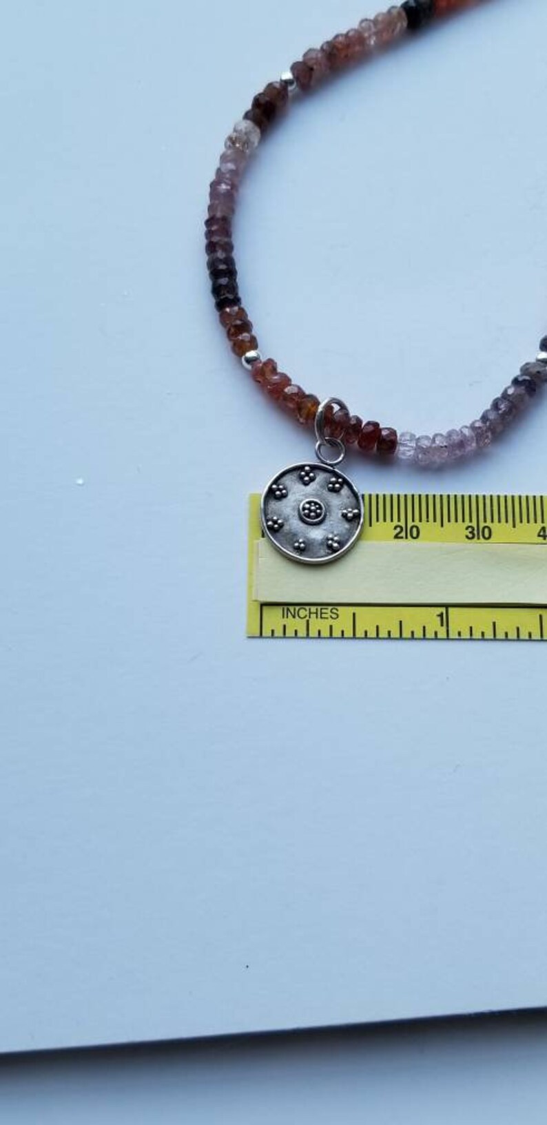 K&ouml;nnte beinhalten: Eine silberne Anh&auml;nger-Kette mit einem runden, silbernen Anh&auml;nger mit einem floralen Design. Die Kette besteht aus kleinen, mehrfarbigen Perlen.
