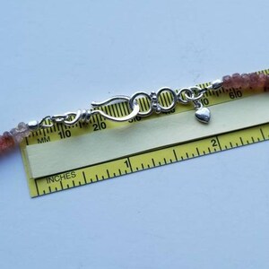 K&ouml;nnte beinhalten: Nahaufnahme einer silbernen Schlie&szlig;e mit einem Herz-Anh&auml;nger an einer Perlenkette. Die Kette wird auf einem Lineal mit Markierungen in Zoll und Millimetern gemessen.