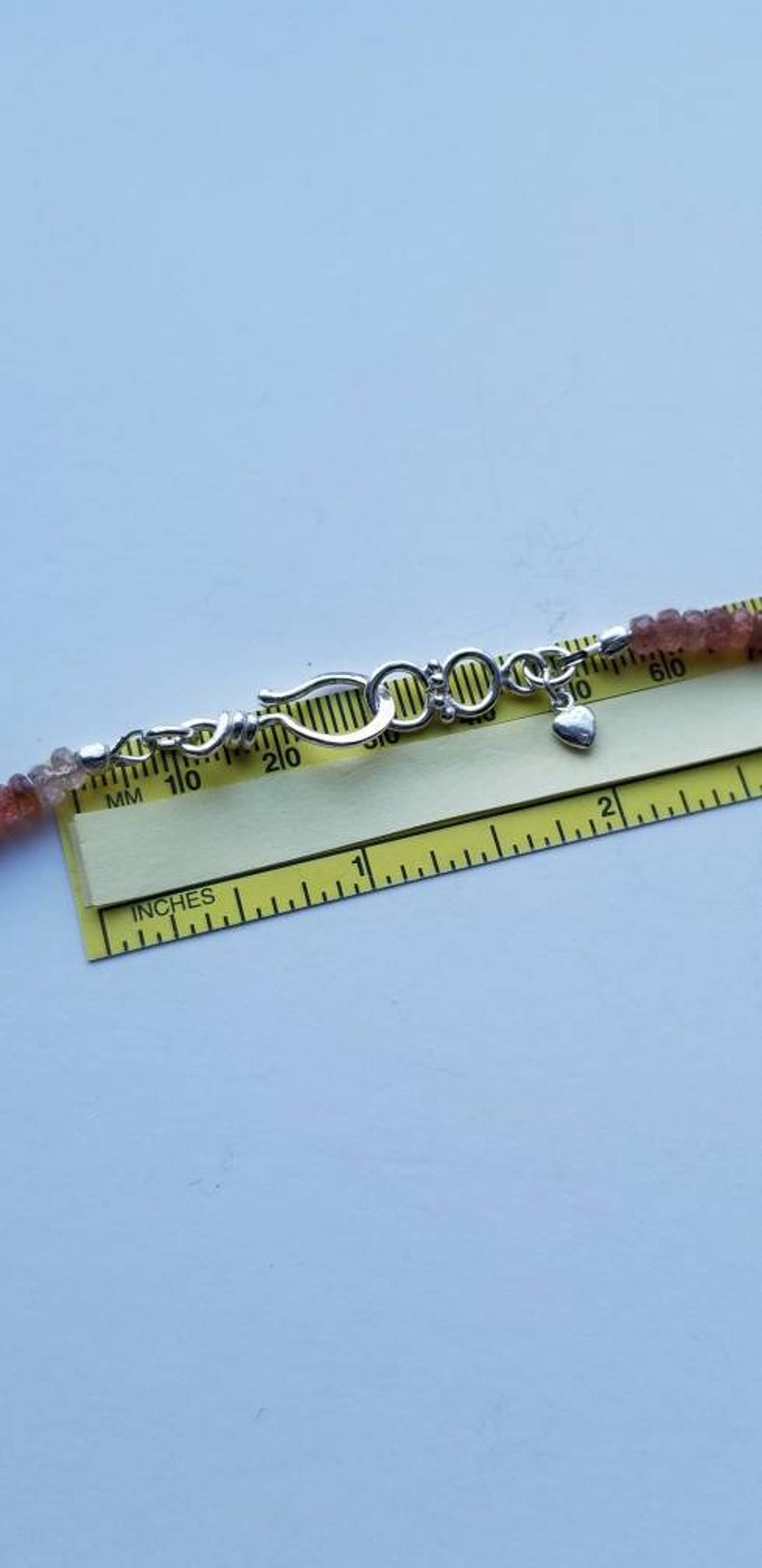 K&ouml;nnte beinhalten: Nahaufnahme einer silbernen Schlie&szlig;e mit einem Herz-Anh&auml;nger an einer Perlenkette. Die Kette wird auf einem Lineal mit Markierungen in Zoll und Millimetern gemessen.
