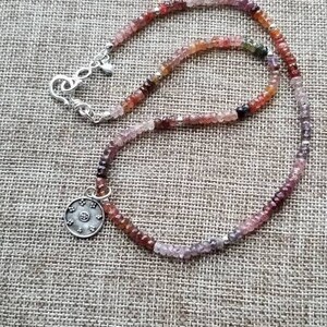 K&ouml;nnte beinhalten: Eine silberne Halskette mit einem runden Anh&auml;nger und einer mehrfarbigen Edelsteinkette. Die Kette besteht aus kleinen, facettierten Edelsteinen in den Farben Rosa, Rot, Lila und Gr&uuml;n.
