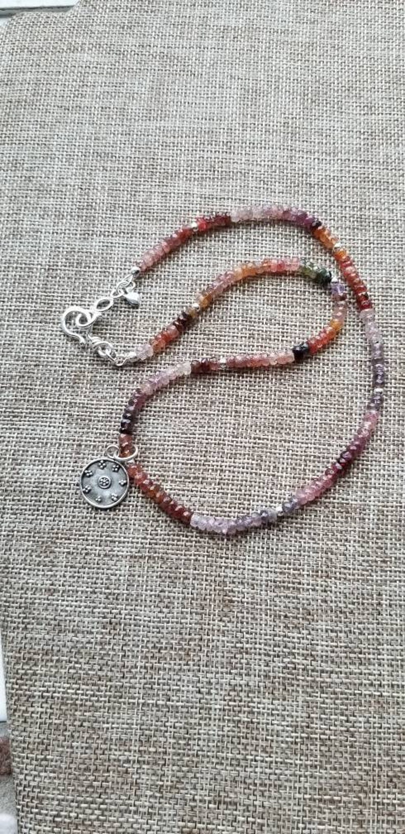 K&ouml;nnte beinhalten: Eine silberne Halskette mit einem runden Anh&auml;nger und einer mehrfarbigen Edelsteinkette. Die Kette besteht aus kleinen, facettierten Edelsteinen in den Farben Rosa, Rot, Lila und Gr&uuml;n.
