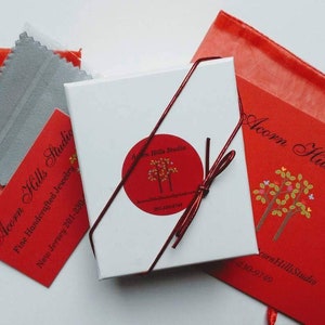 K&ouml;nnte beinhalten: Eine wei&szlig;e Geschenkbox mit einem roten Band, das darum gebunden ist. Die Box hat einen roten und wei&szlig;en Aufkleber mit dem Text "Acorn Hills Studio" und einem kleinen Bild von zwei B&auml;umen. Die Box steht auf einer wei&szlig;en Oberfl&auml;che neben einer roten Geschenkt&uuml;te und einem kleinen grauen Poliertuch. Die Geschenkt&uuml;te hat den Text "Acorn Hills Studio" aufgedruckt.