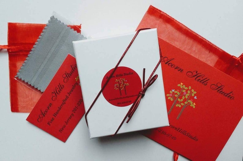 K&ouml;nnte beinhalten: Eine wei&szlig;e Geschenkbox mit einem roten Band, das darum gebunden ist. Die Box hat einen roten und wei&szlig;en Aufkleber mit dem Text "Acorn Hills Studio" und einem kleinen Bild von zwei B&auml;umen. Die Box steht auf einer wei&szlig;en Oberfl&auml;che neben einer roten Geschenkt&uuml;te und einem kleinen grauen Poliertuch. Die Geschenkt&uuml;te hat den Text "Acorn Hills Studio" aufgedruckt.