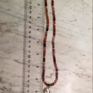 K&ouml;nnte beinhalten: Eine silberne Halskette mit einem kleinen runden Anh&auml;nger mit einem Blumenmuster. Die Halskette besteht aus kleinen, mehrfarbigen Perlen in den Farben Rosa, Rot und Braun.