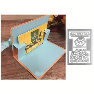 May include: A blue card holder with a gift card inside. The card holder has a pop-up feature and is decorated with a brown and orange pattern. The gift card is from "Taverna Bakery" and says "Gift Card". A silver die-cut card with a floral design and the word "Thanks" is also shown.