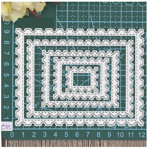 May include: A set of five white metal die-cut frames with scalloped edges. The frames are nested inside each other, creating a layered effect. The frames are measured in centimeters on a green cutting mat.