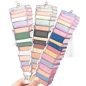 以下が含まれることがあります： 花柄のアクセントがついた本が積み重ねられた、カラフルなブックマーク3つ。各ブックマークにはタッセルが付いています。