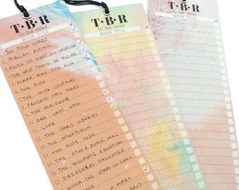 Puede incluir: Tres marcadores coloridos con un fondo de acuarela y una lista de títulos de libros para leer. Los marcadores tienen una cinta negra en la parte superior y el texto "T-B-R to be read BOOK TITLE" en la parte superior.