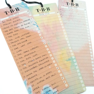 May include: Three colorful bookmarks with a watercolor background and a list of book titles to be read. The bookmarks have a black ribbon at the top and the text "T-B-R to be read BOOK TITLE" at the top.