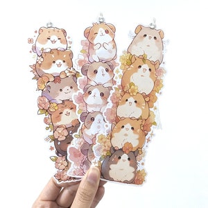 May include: A set of 10 clear acrylic bookmarks featuring cute cartoon hamsters in various poses with floral accents. The bookmarks are stacked in a W shape and are held in a hand.