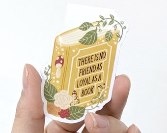 Puede incluir: Una pegatina amarilla con forma de libro con el texto "THERE IS NO FRIEND AS LOYAL AS A BOOK". La pegatina está decorada con ilustraciones florales y de setas. La pegatina está sujeta por una mano.
