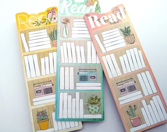 Puede incluir: Tres marcadores de colores con un diseño de estantería. Cada marcador tiene un esquema de color diferente y presenta una pequeña planta en maceta, una radio vintage y la palabra "Leer" en la parte superior.