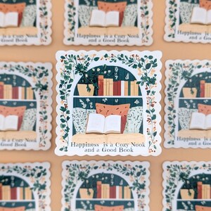 Puede incluir: Una pegatina que representa un rinc&oacute;n de lectura acogedor. El dise&ntilde;o incluye una estanter&iacute;a, un libro abierto, almohadas y plantas, enmarcado por un borde floral. Se muestra el texto "Happiness is a Cozy Nook and a Good Book".