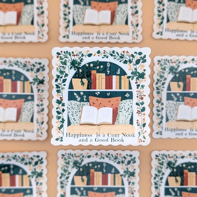 Puede incluir: Una pegatina que representa un rinc&oacute;n de lectura acogedor. El dise&ntilde;o incluye una estanter&iacute;a, un libro abierto, almohadas y plantas, enmarcado por un borde floral. Se muestra el texto "Happiness is a Cozy Nook and a Good Book".