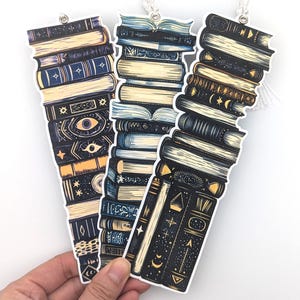 Peut inclure: Trois marque-pages décoratifs représentant des piles de livres avec des motifs célestes et mystiques. Les marque-pages ont un pompon blanc et un anneau en métal argenté. La palette de couleurs comprend le bleu marine, l'or et la crème.