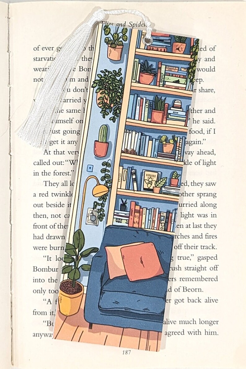 Pode incluir: Um marcador de p&aacute;gina com uma ilustra&ccedil;&atilde;o colorida de uma estante com livros, plantas e uma poltrona aconchegante. O marcador &eacute; feito de acr&iacute;lico transparente e tem uma borla branca.